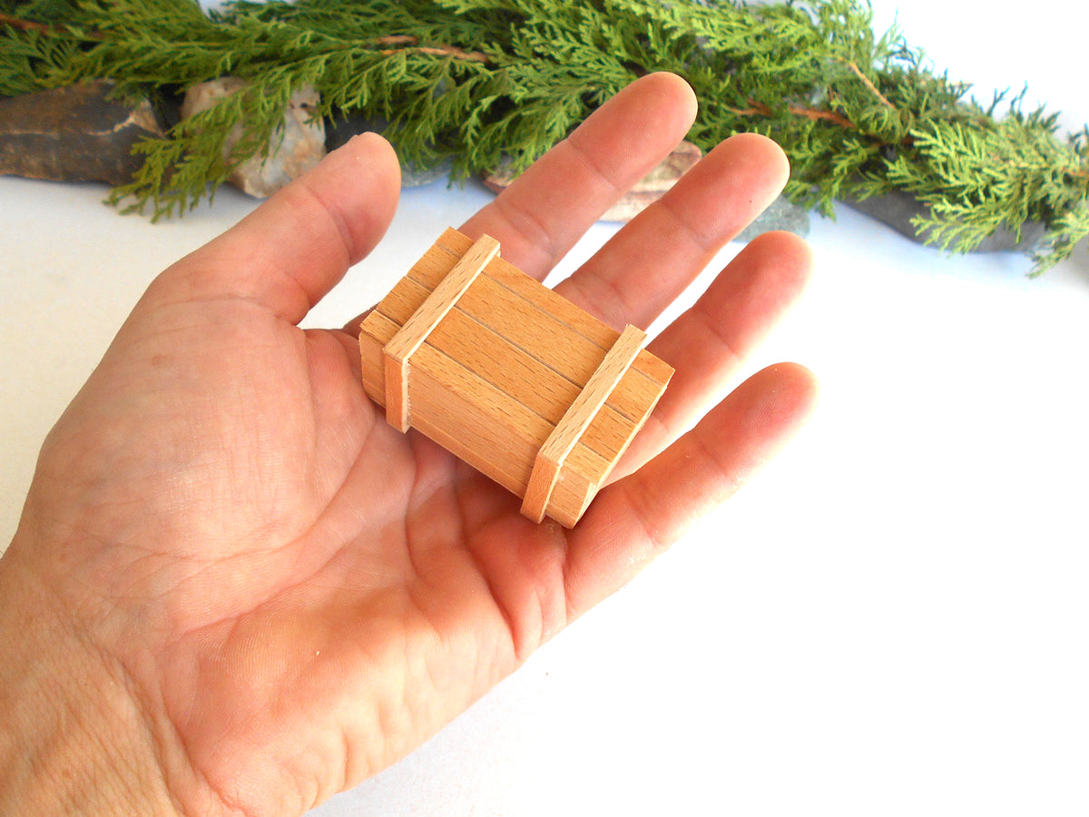 A miniature wooden box with a light natural wood finish, held in the palm of a hand to show its small scale, with green foliage in the background.