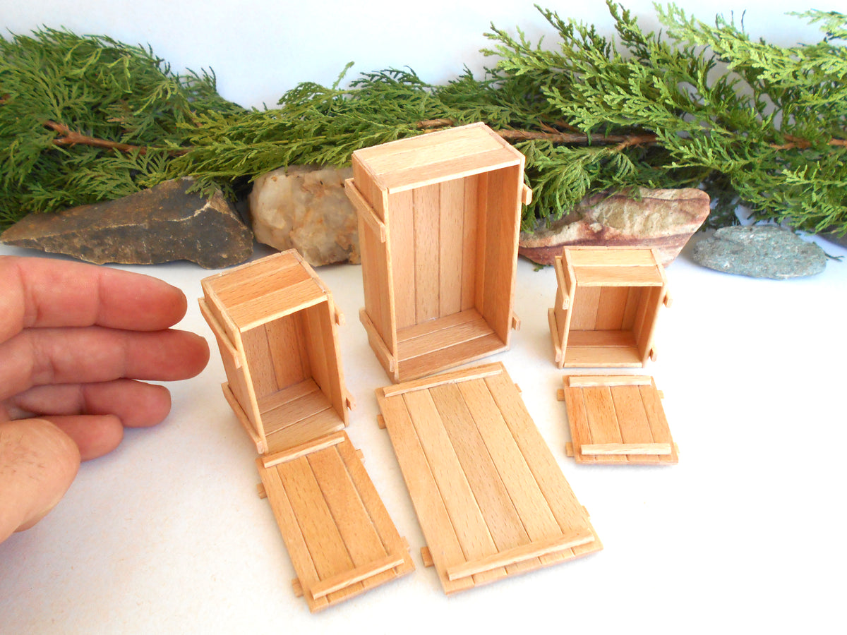 Five miniature light wood crate and platform pieces arranged on a white surface, with a hand visible on the left for scale and greenery/rocks in the background.