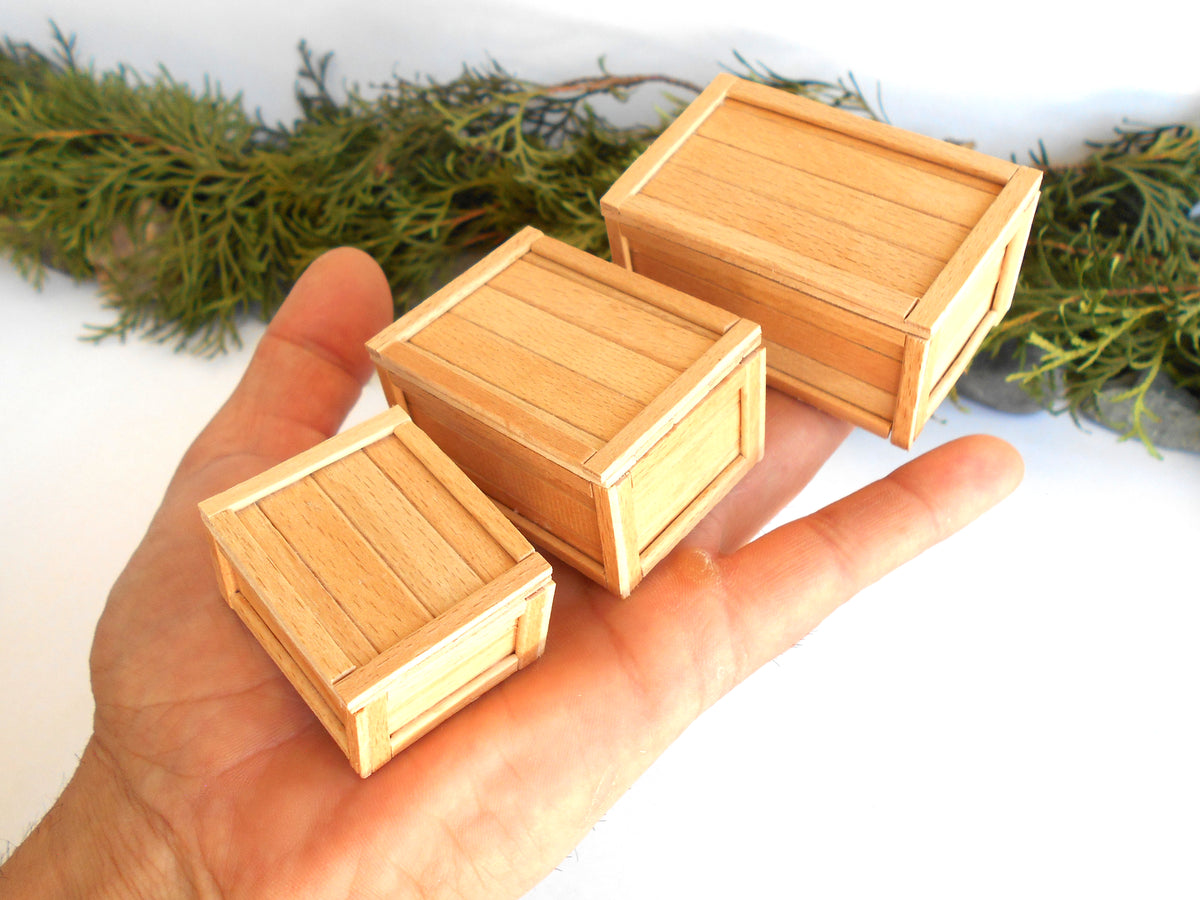 Three miniature light wood crates of different sizes held in a person's hand, with green pine branches in the background.