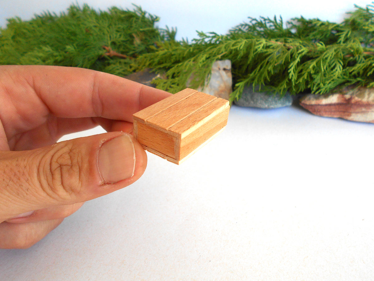 A hand holding a small, light-colored wooden box with a herringbone pattern, set against a white background with green foliage and stones in the blurred background.