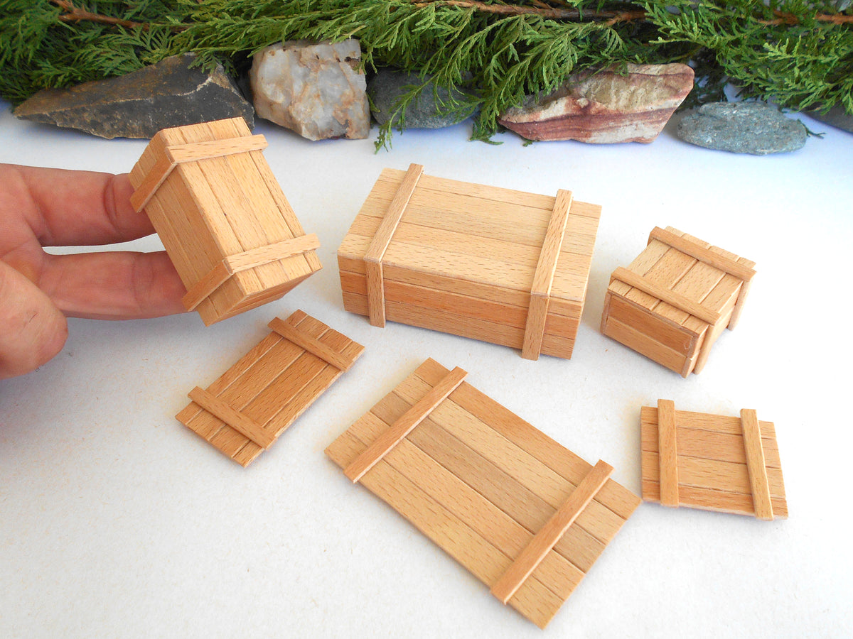 Six miniature wooden crates in various sizes, including one being held by a hand, displayed on a white surface with a natural stone and pine background.