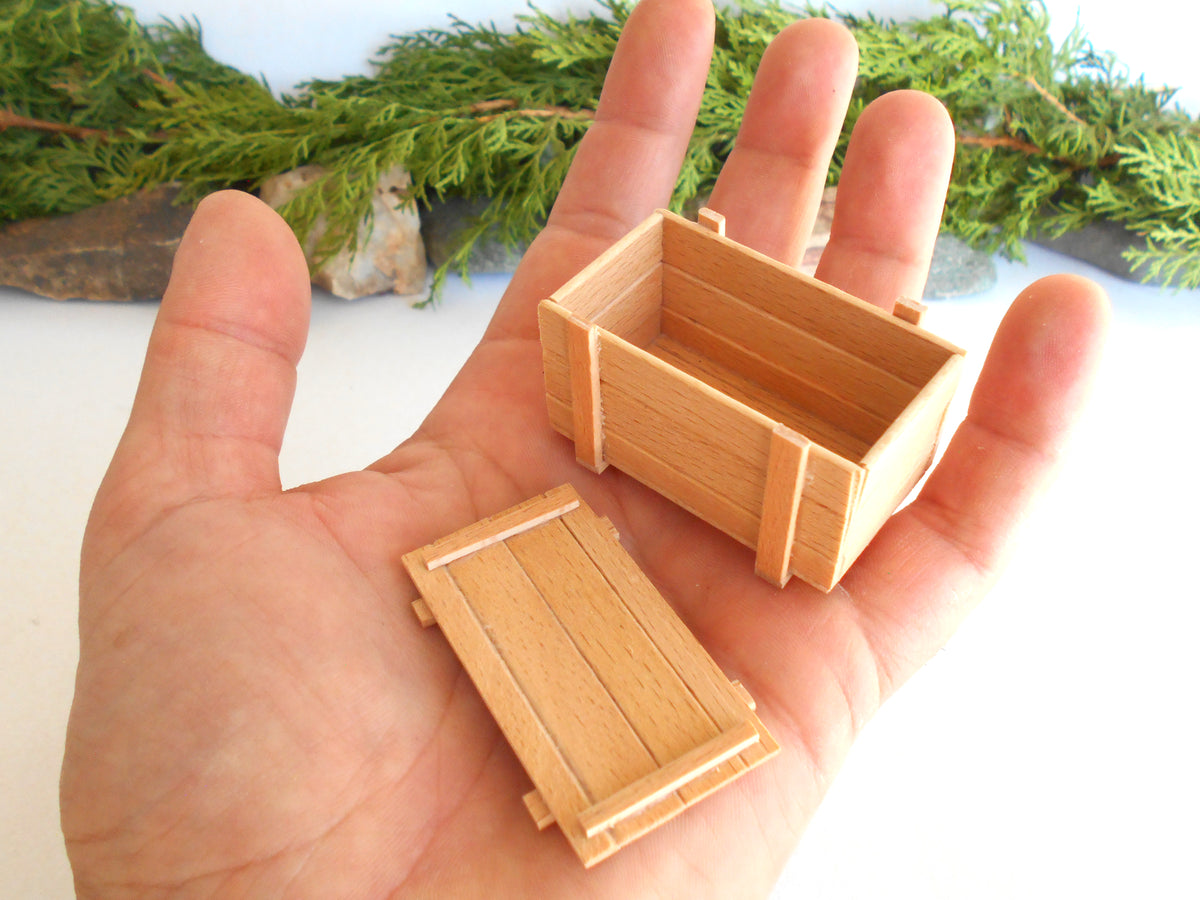 A miniature wooden crate box and its matching lid held in a person's hand, with green foliage in the background.