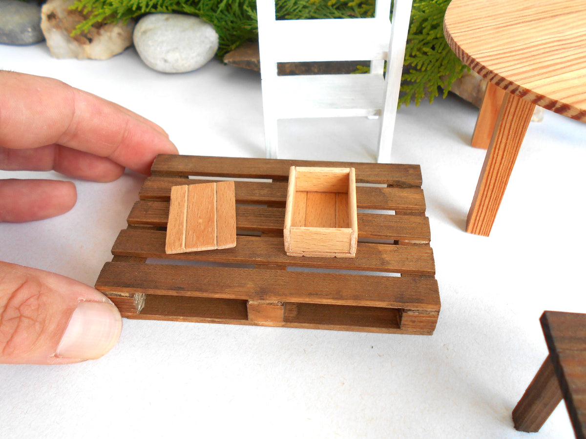 A miniature wooden pallet table accessory set held by a hand, featuring a brown wooden slat base with a small rectangular crate and a separate matching lid piece, shown in a miniature-scale scene.