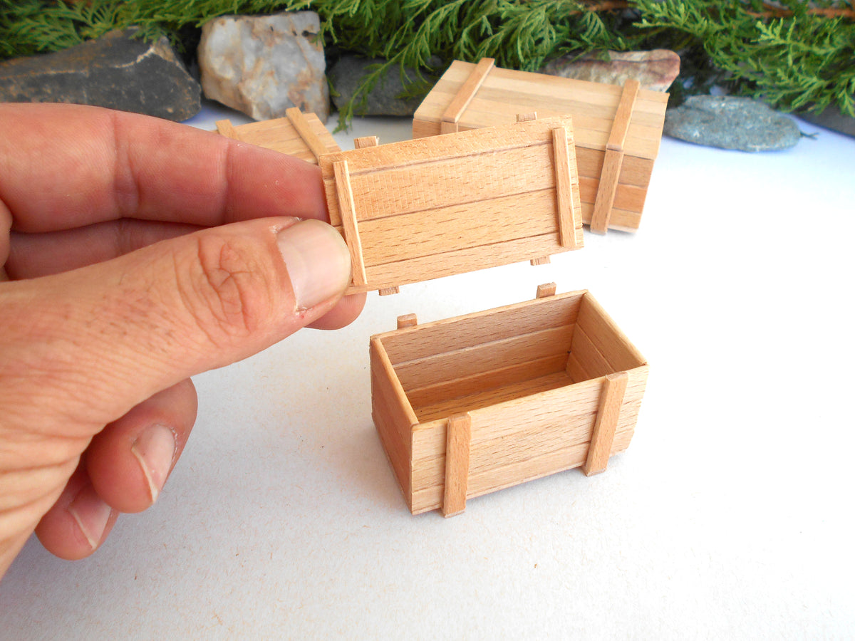 A hand holding the lid of a small wooden crate box, with two more matching wooden crate boxes on a white surface in the background.
