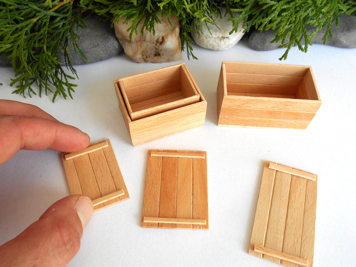 A hand is holding a miniature wooden crate lid next to several matching miniature wooden storage crates on a white surface with decorative greenery in the background.
