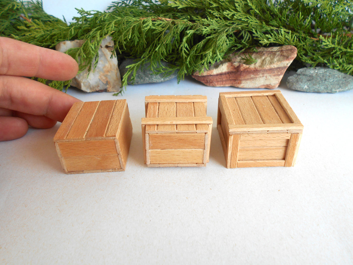 Three miniature light brown wooden crates of different sizes arranged on a white surface, with a person's hand pointing to the leftmost crate and green foliage in the background.