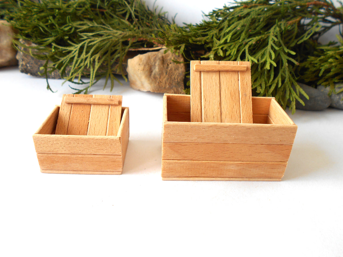 Two miniature light wood boxes with slatted lids, shown on a white surface with green foliage and small stones in the background.