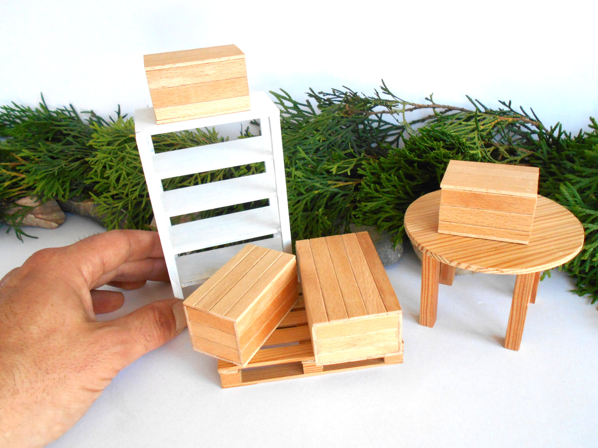 A hand is holding a miniature wooden pallet toy next to other dollhouse furniture pieces, including a white pallet rack, a round wooden table with a wooden crate, and several small wooden blocks, set against a background of green pine garland and a white wall.