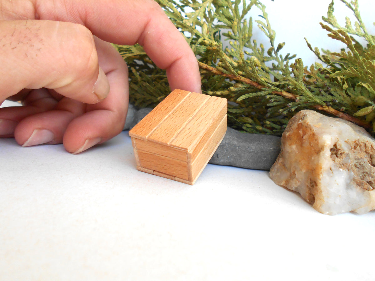 A miniature light brown wooden box with visible wood grain, held by a human hand next to a gray stone and a natural rock on a white surface.
