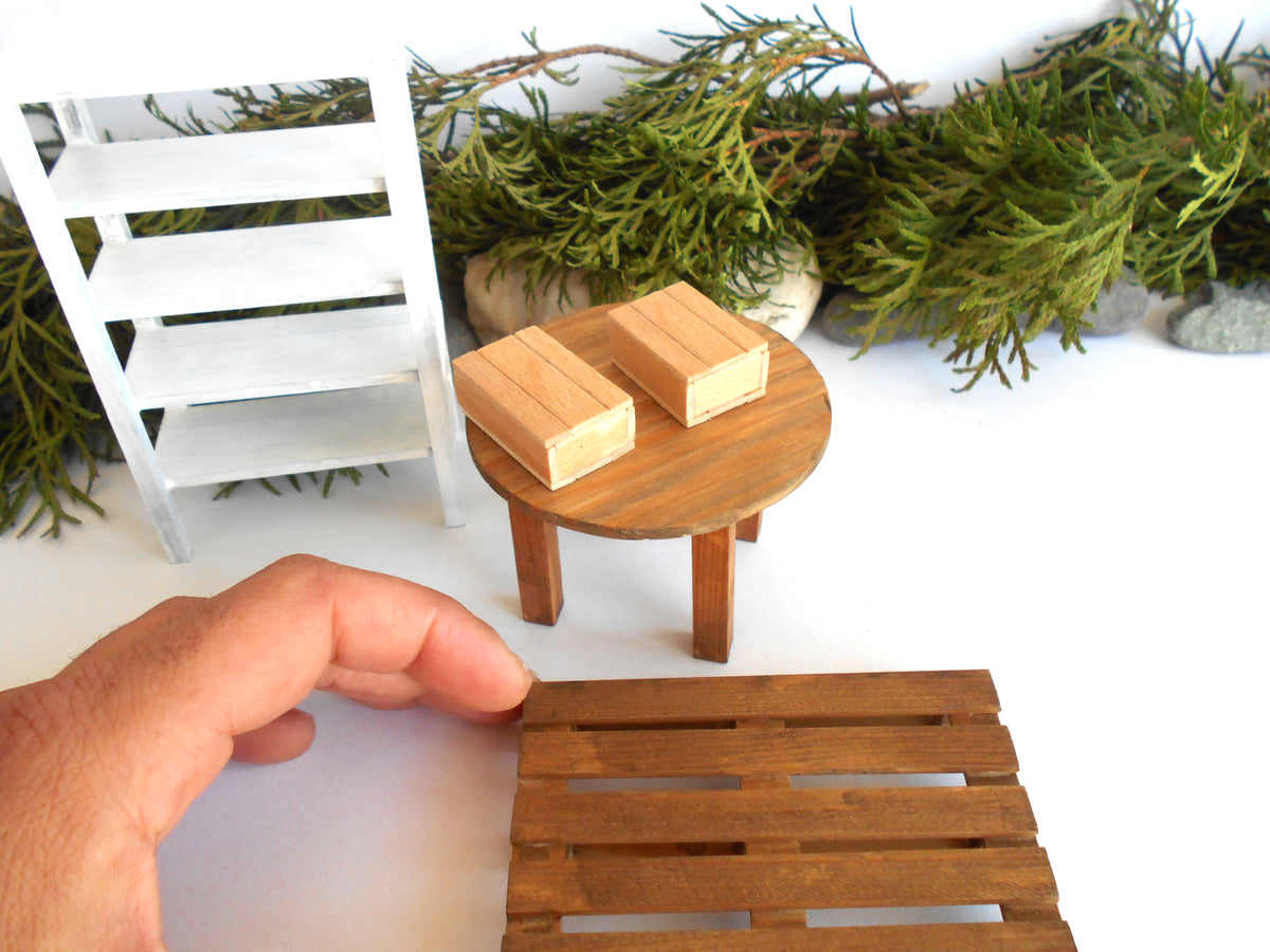 A miniature wooden table set with a round brown table, two small wooden blocks, and a hand holding a matching miniature wooden slatted stool in the foreground. The scene is styled with a white miniature shelf and green foliage in the background.