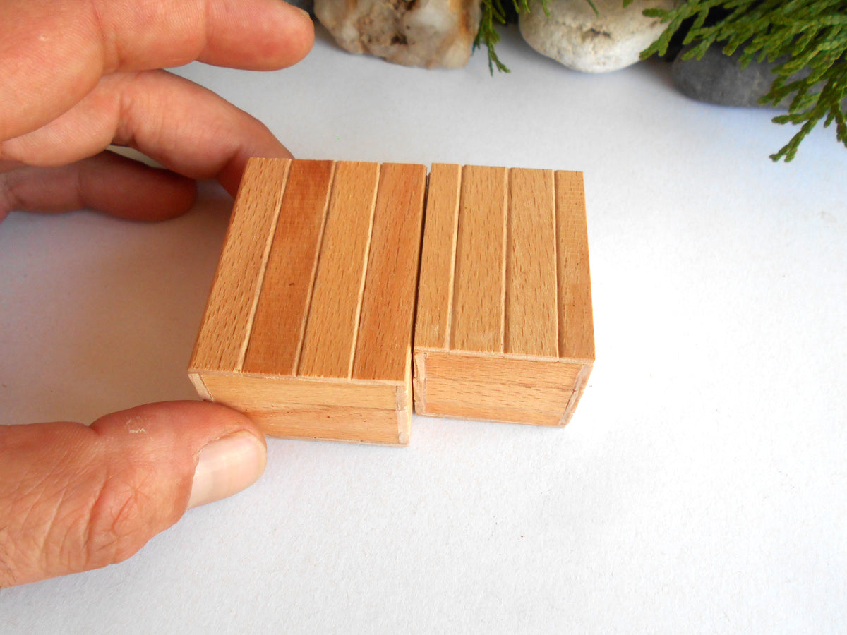 Two small rectangular wooden blocks with vertical grooves, held by a person's hand on a white surface with blurred decorative rocks and greenery in the background.