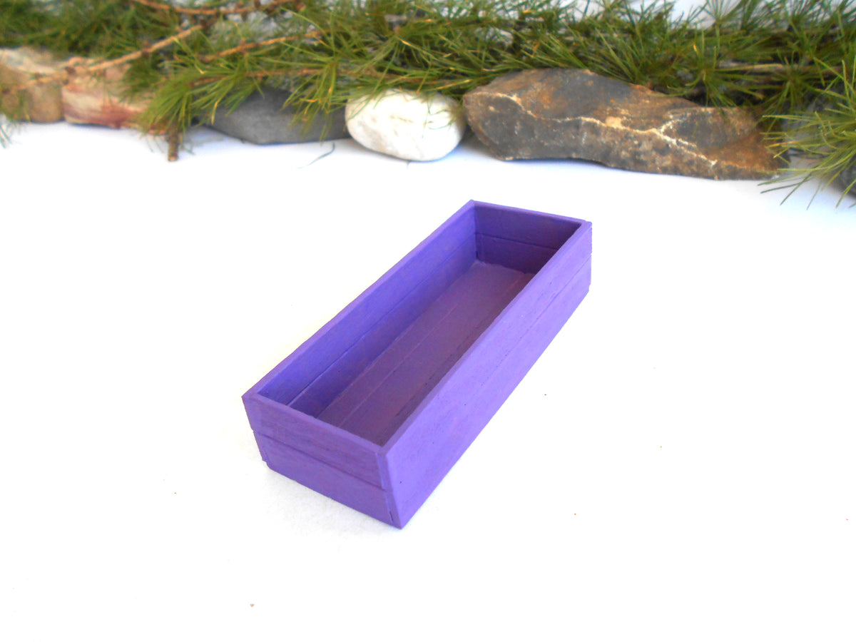 A purple rectangular wooden box with an open top, photographed on a white surface with pine branches and rocks in the background.