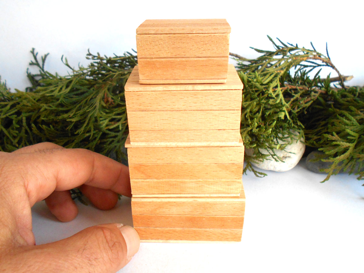 A hand holds a stack of four miniature, light-colored wooden boxes with a natural oak finish, set against a white background with green foliage.
