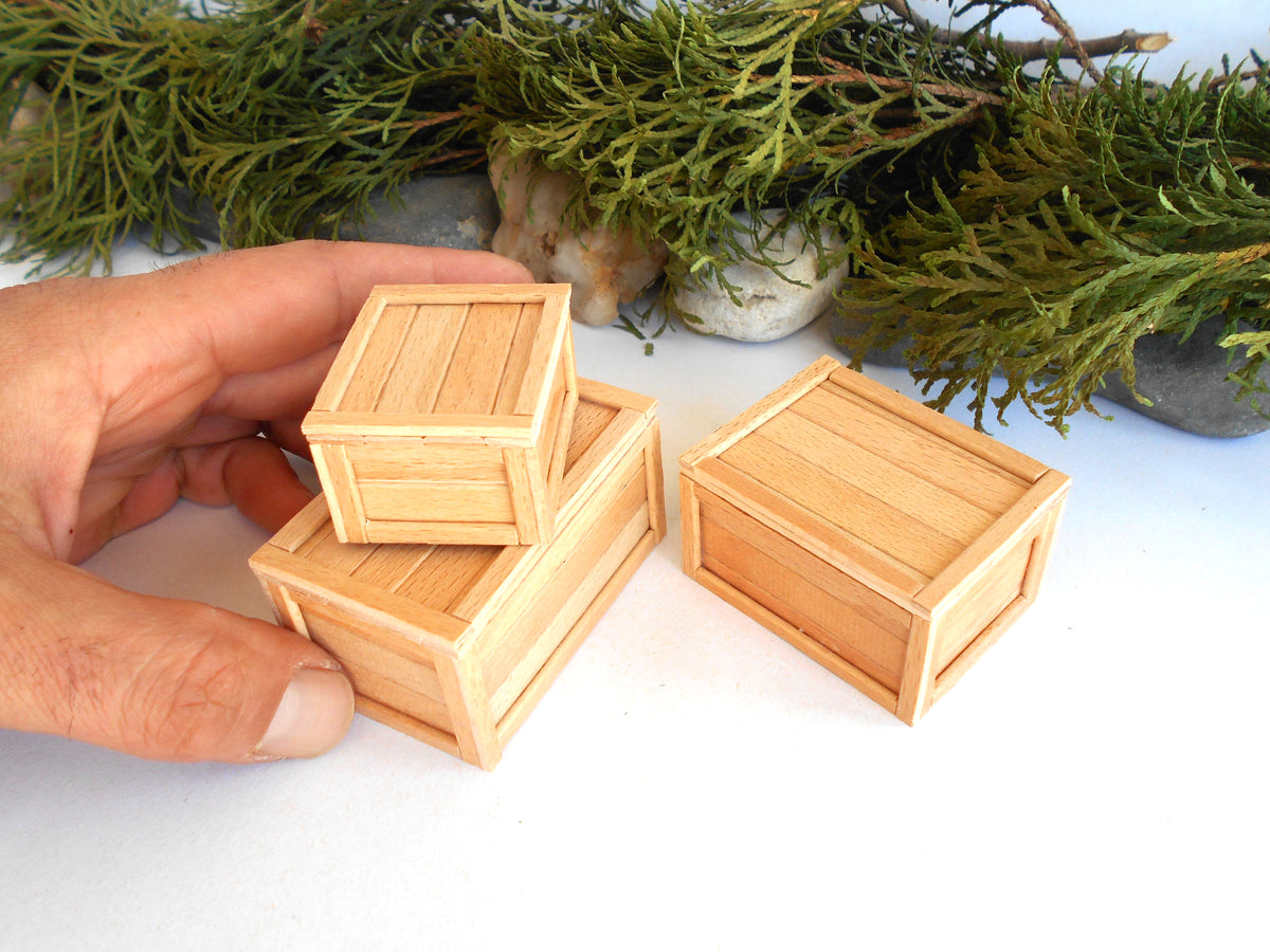 Three miniature light wood crates, including a stack of two held by a hand and one standing beside it, with green foliage in the background.