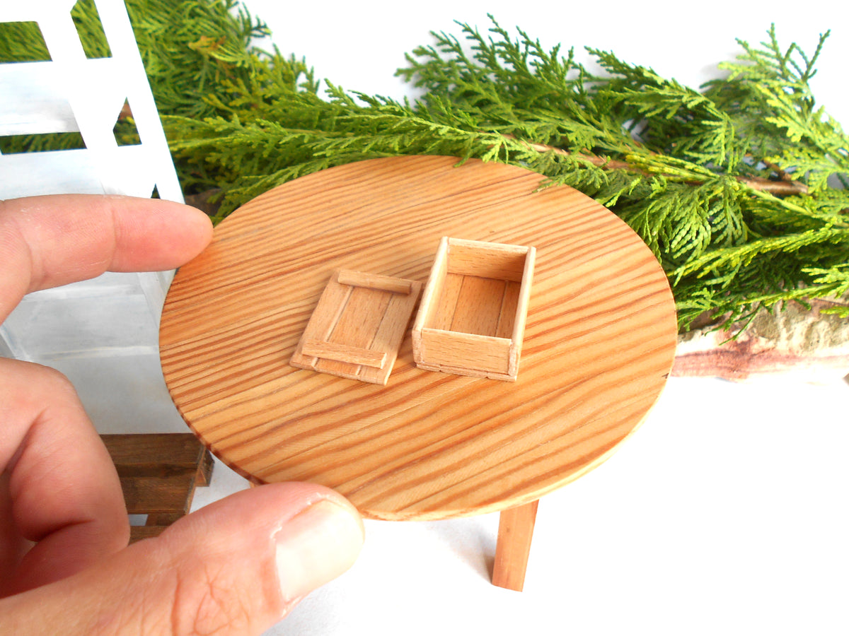 A miniature round wooden table with two small wooden boxes on top, held by a hand to show scale, with green foliage and a white background.