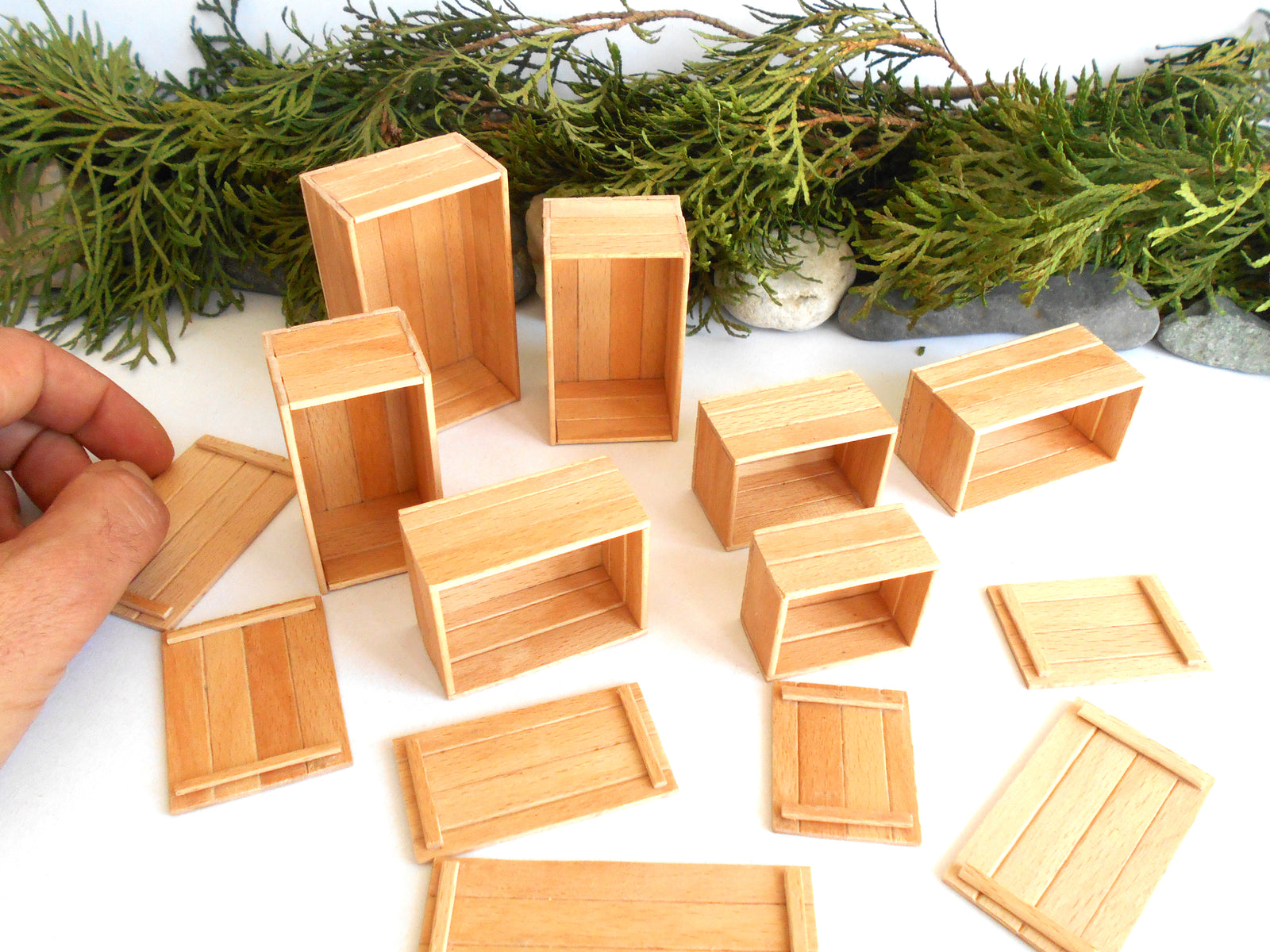 A set of miniature light wood crates and boxes, including some lids, displayed on a white surface with a green pine garland in the background. A hand is visible on the left holding one of the crates.