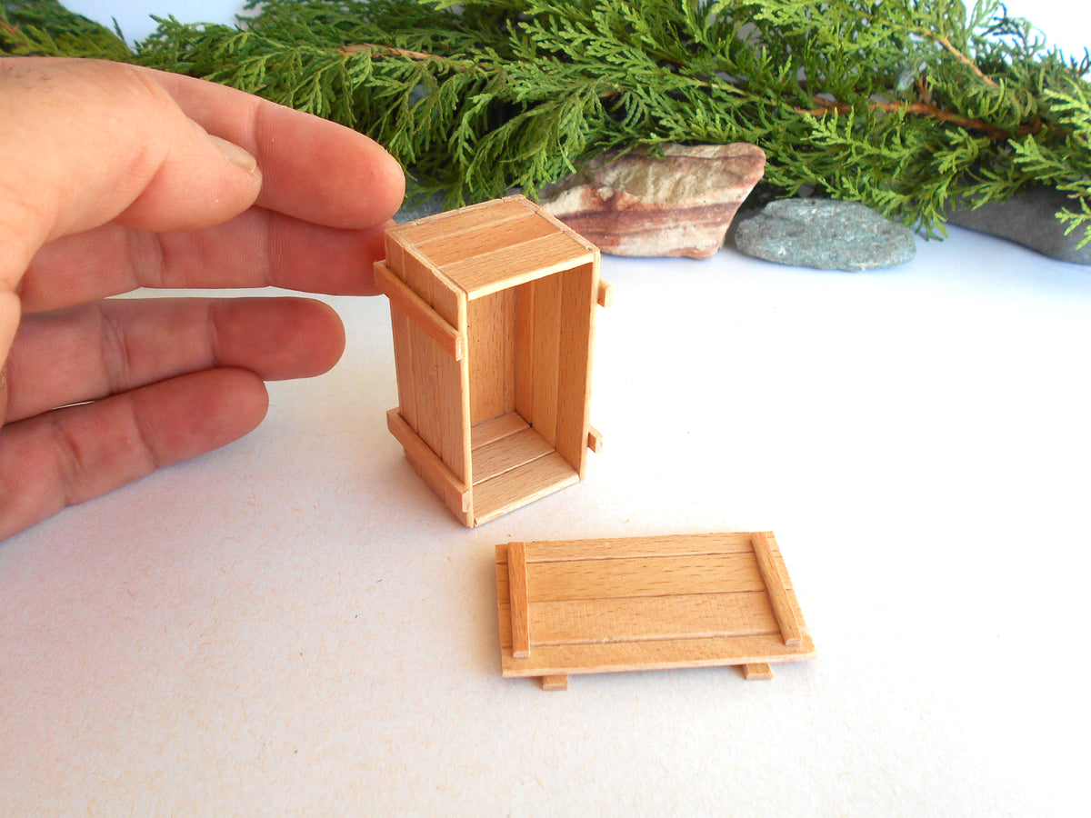 A miniature wooden box with its lid placed beside it on a white surface, with a person's hand holding the box and evergreen branches in the background.