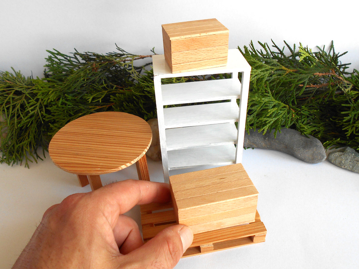 A hand holds a miniature wooden box on a small wooden base in front of a white slatted cabinet, with a round wooden stool and green foliage in the background.