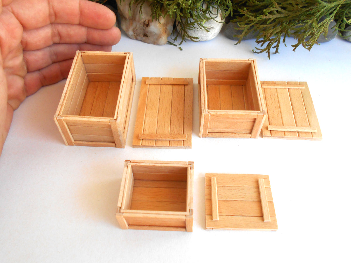 Five miniature light wood crates of different sizes and configurations, shown with their lids beside them on a white surface, with a hand and green plants in the background for scale and context.