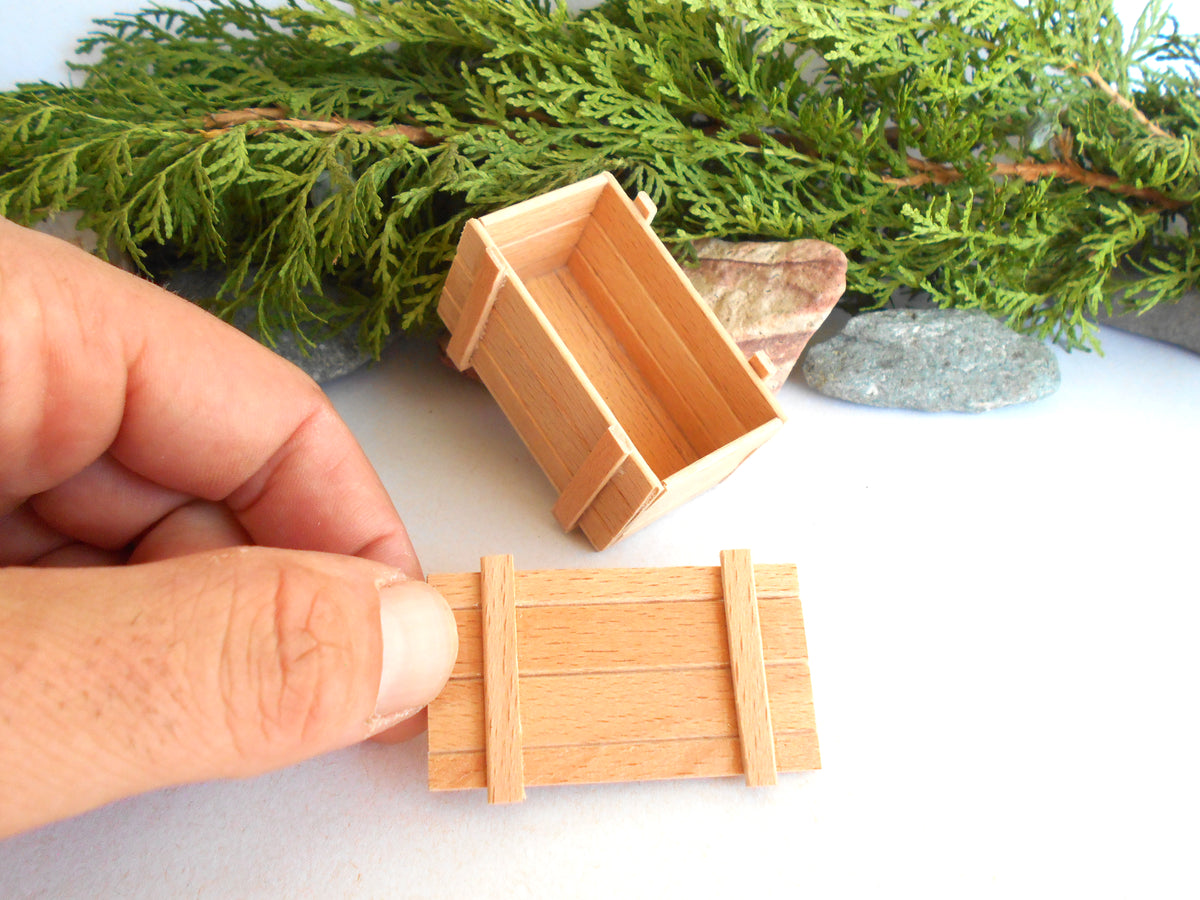 A miniature wooden crate or box shown in two sizes, with one held by a hand and one placed on a white surface with green foliage and small stones in the background.