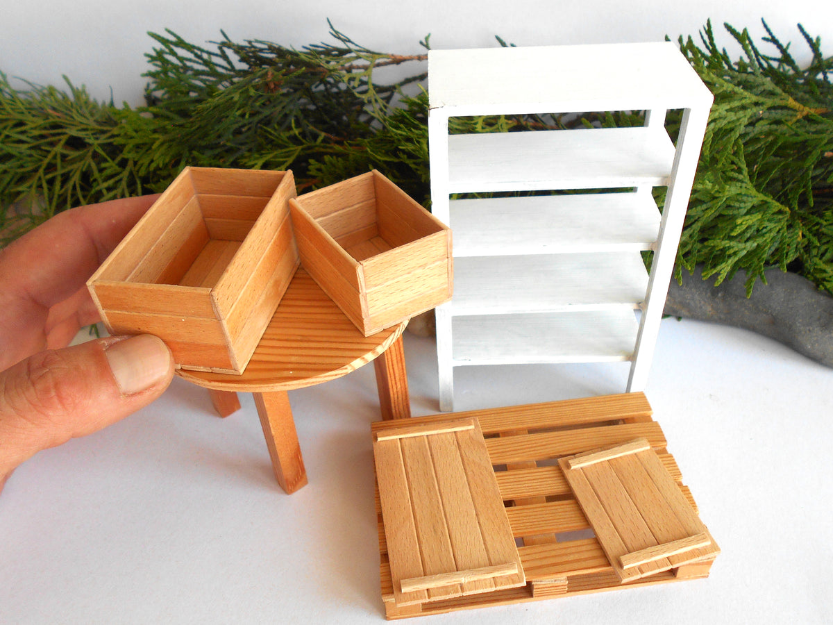 A miniature wooden display stand and crate set shown on a white surface, with a hand holding one of the small wooden crates and a white shelving unit in the background.