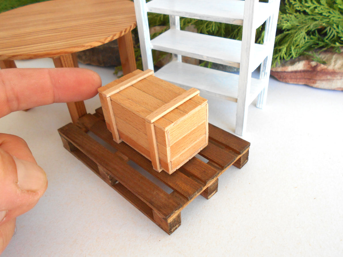 A miniature wooden crate model displayed on a small wooden pallet base, with a human finger pointing at it for scale. The background features a miniature white shelf and a wooden table.