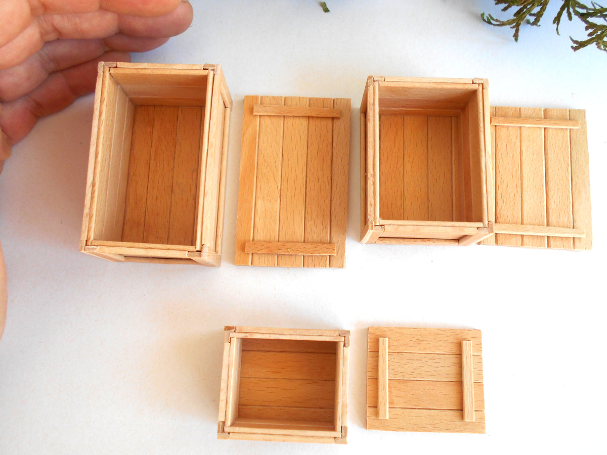 Five miniature light wood crates of different sizes and orientations arranged on a white surface, with a hand holding one crate at the upper left.