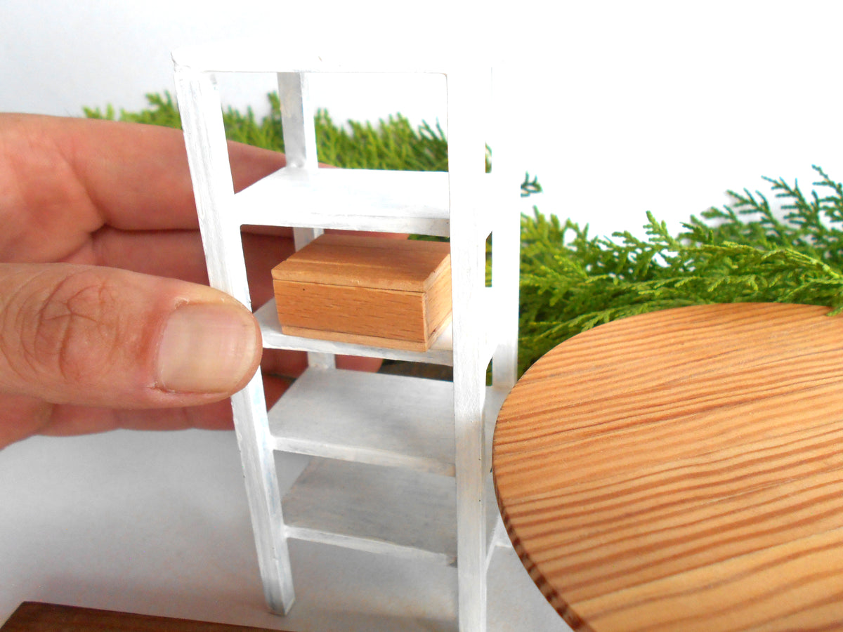 A miniature white shelving unit with a small wooden box on one of its shelves, held by a person's hand to show scale, with a wooden table and green foliage in the background.