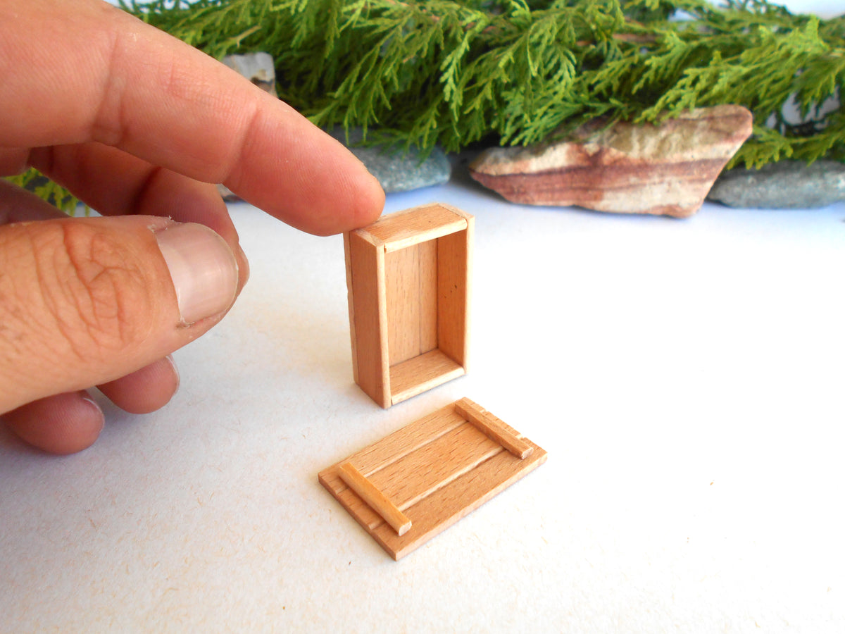 A close-up of a hand holding a miniature light wood box, with a matching small wooden tray or lid piece placed below it on a white surface. The background includes soft-focus green foliage and stones.