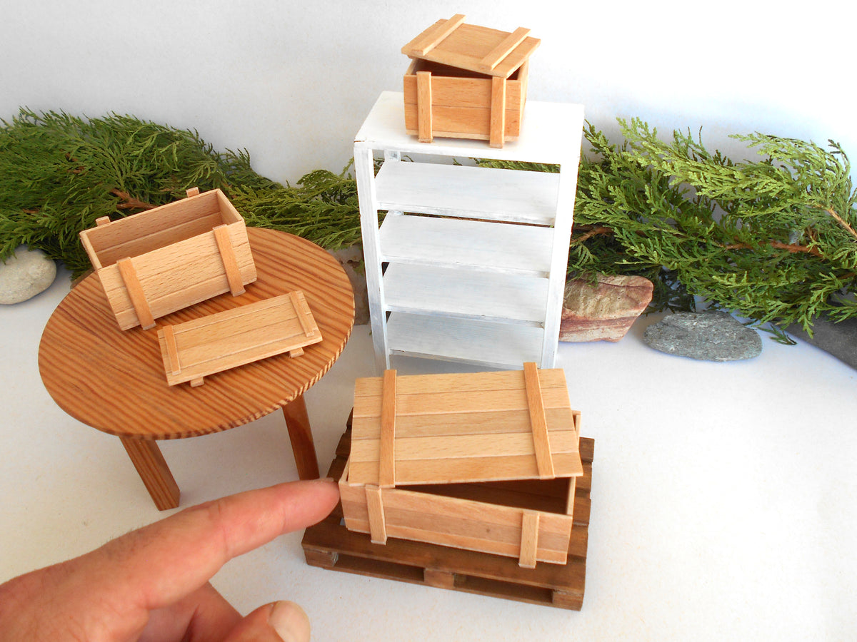 A miniature wooden crate set displayed on a tabletop with a white cabinet, green foliage props, and a person's finger pointing toward the set.