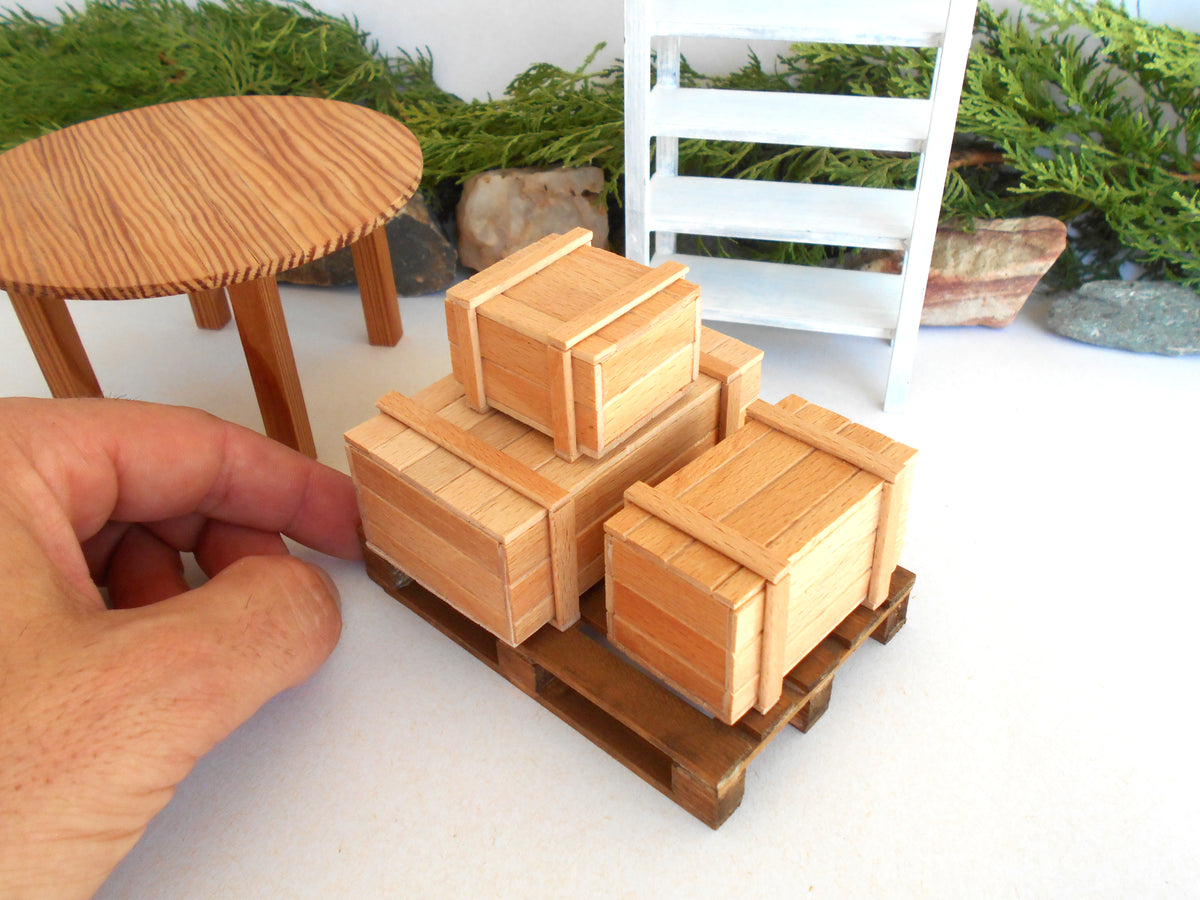 A miniature wooden crate set displayed on a small wooden pallet, with a hand holding the scale reference in the foreground and miniature furniture in the background.
