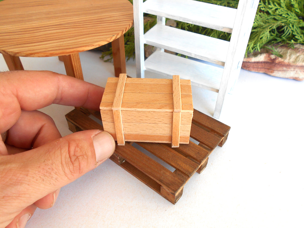 A miniature wooden storage box with two straps, held by a person's hand to show its small scale, set on a miniature wooden pallet in front of a white miniature shelf.