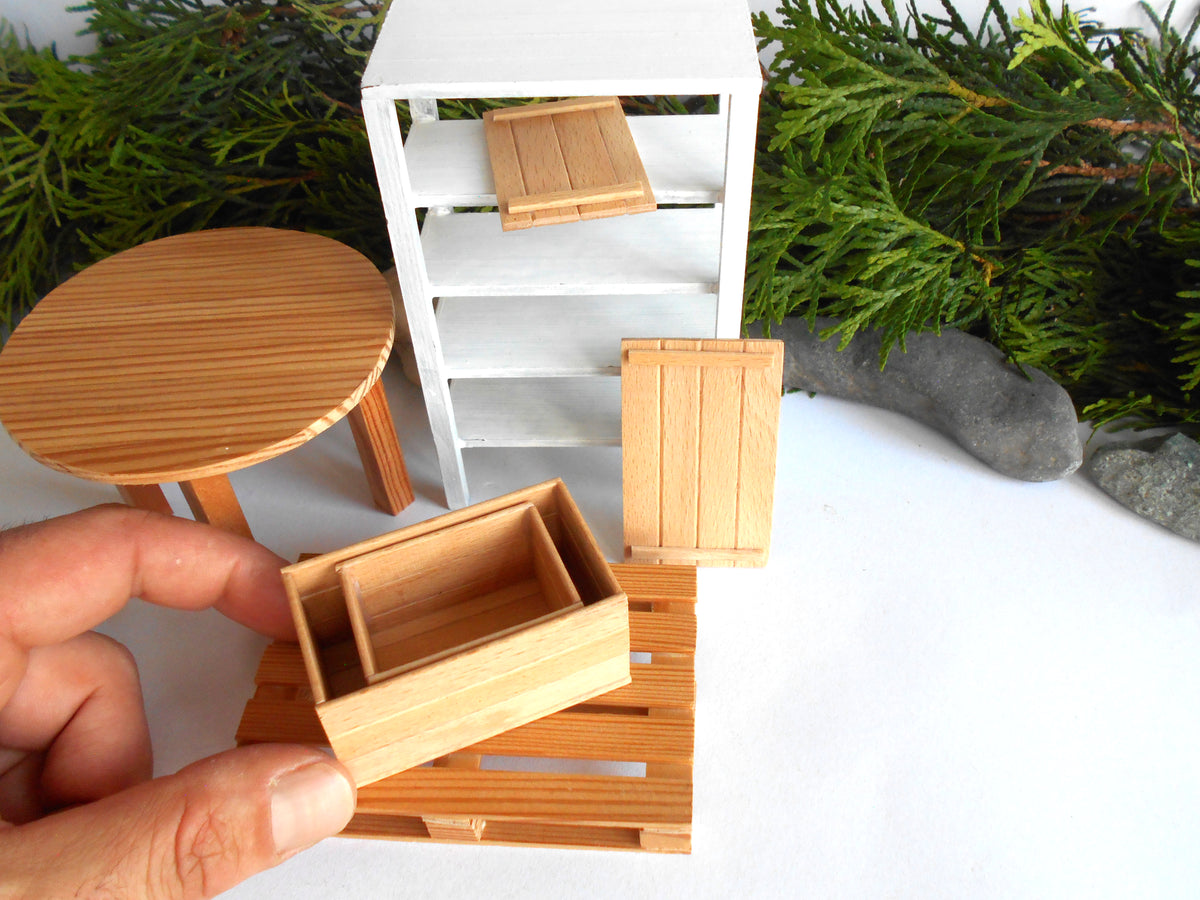 A hand holds a miniature wooden storage box in front of a white shelving unit, with a round wooden table and several small wooden furniture pieces arranged on a white surface with green foliage in the background.