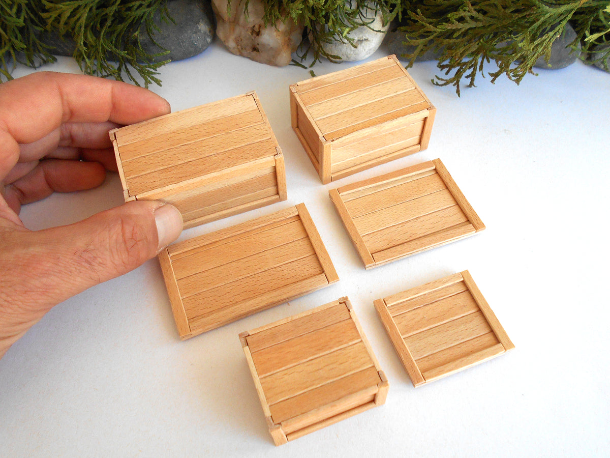A hand holding a miniature wooden crate next to five other small wooden crates and boxes on a white surface with green foliage in the background.