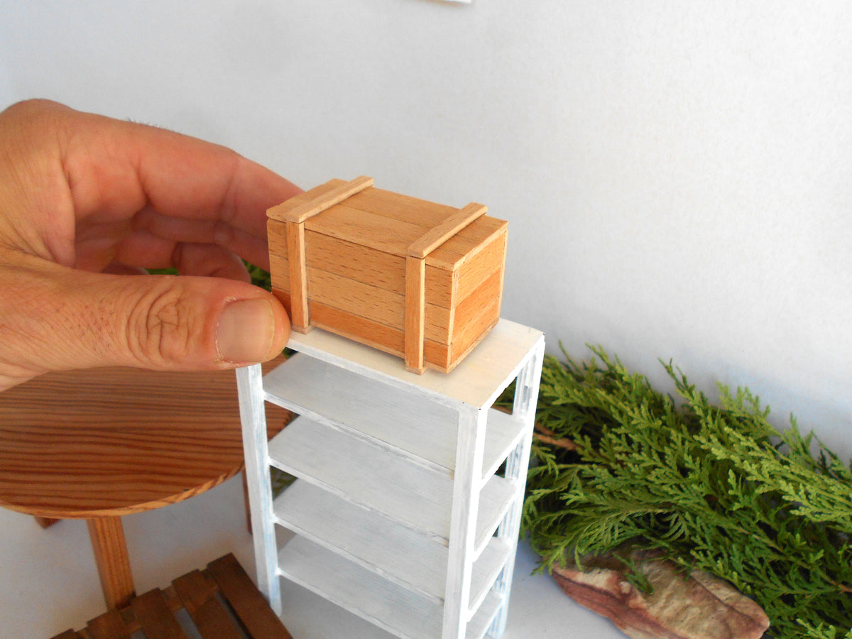 A miniature wooden storage box with a hinged lid, held by a hand on top of a small white shelf. The miniature setup is photographed in a real environment with a white wall, green foliage, and a wooden table visible.