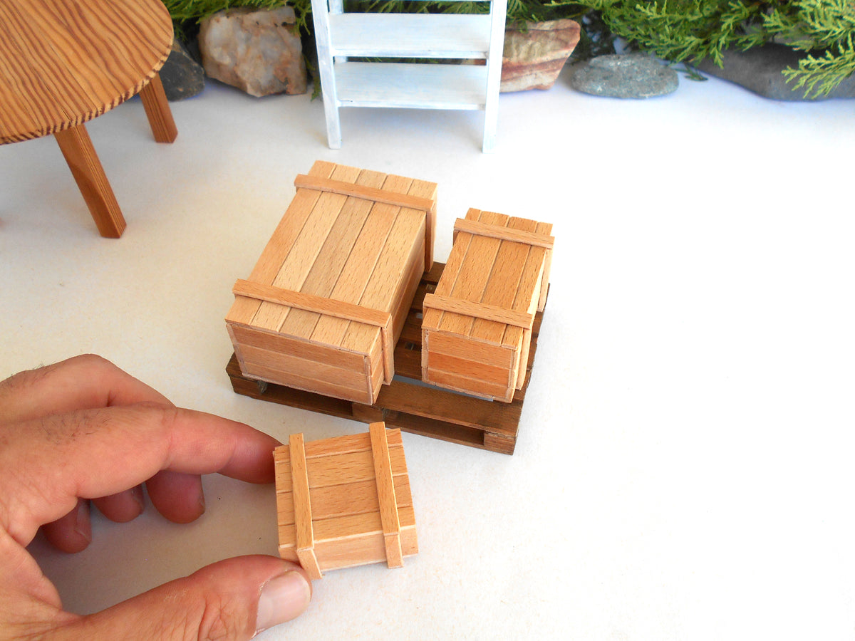 A hand holds a small wooden crate while two larger miniature wooden crates rest on a miniature trailer, with a miniature table and other scale props in the background.