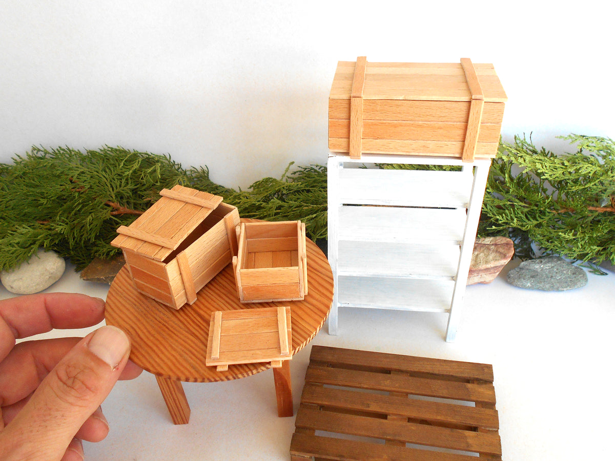 A miniature wooden dollhouse furniture set including a round table with small wooden crates, a white shelving unit with a wooden crate on top, and a wooden pallet, shown with a hand holding one of the crates.