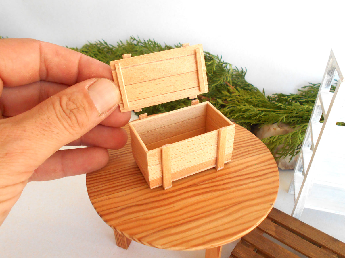 A miniature wooden storage box with its lid open, held by a hand over a small round wooden table. The background features green foliage and a white shelf.
