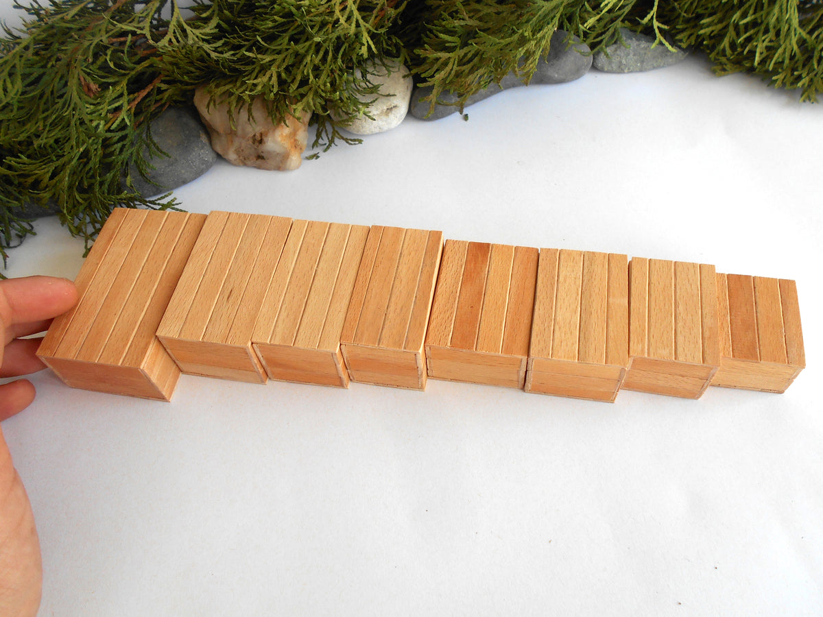A hand holding a row of small natural wood boxes or tiles on a white surface, with greenery and stones in the background.