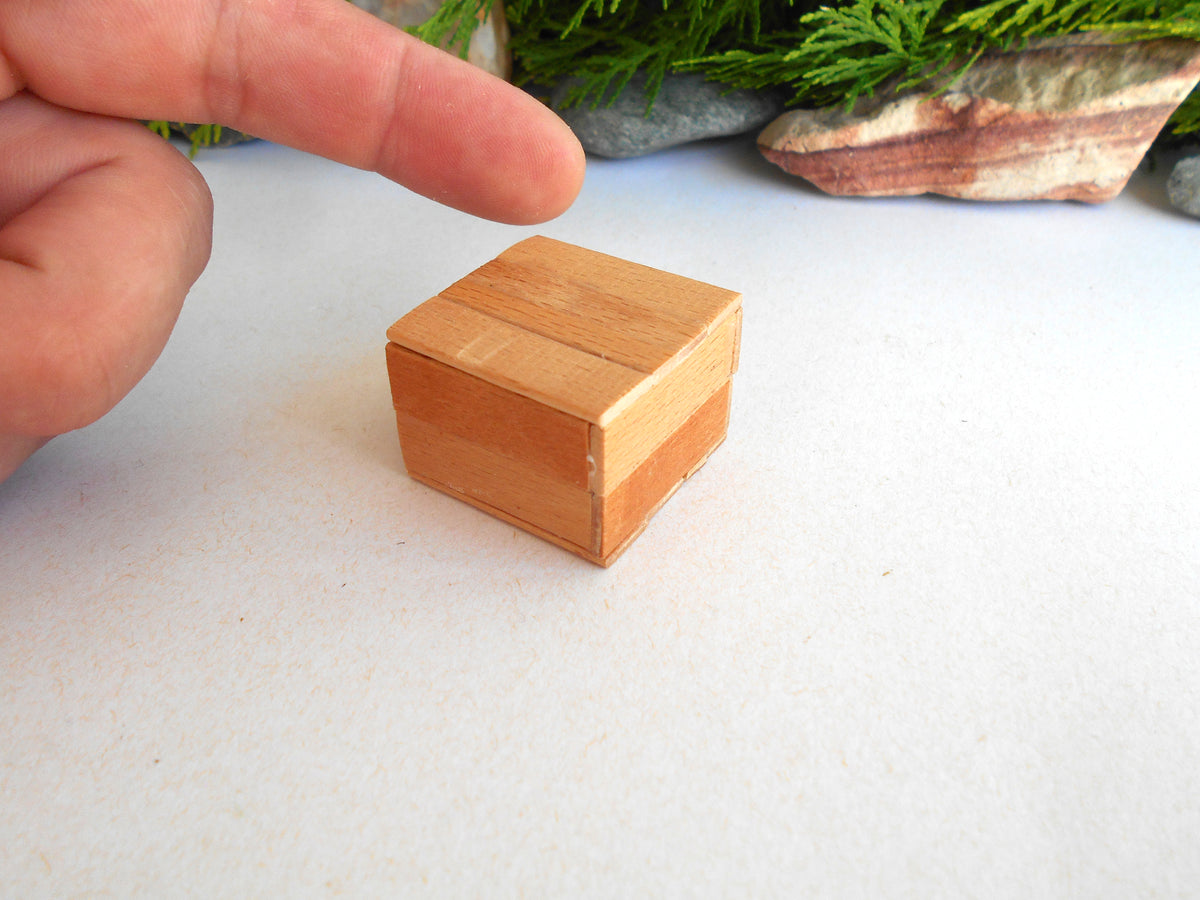 A small square wooden box with a light natural wood finish sits on a white surface, with a person's finger pointing at it and blurred greenery in the background.