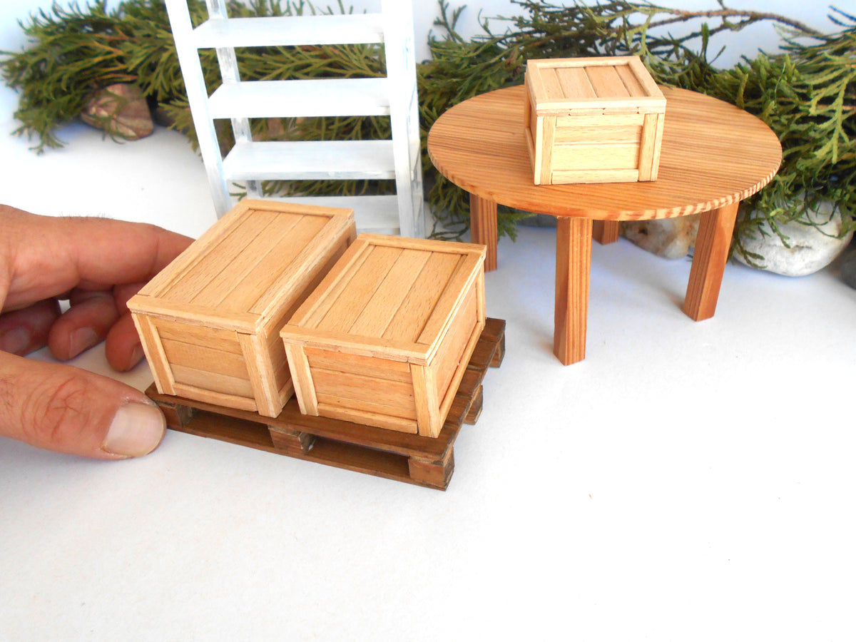 A miniature wooden crate set for dollhouse scale, featuring two small light wood crates on a miniature pallet being held by a hand, with a round wooden table and another crate in the background.