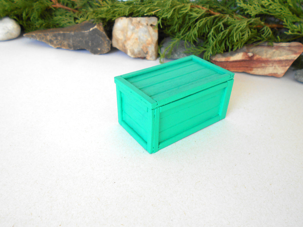 A miniature bright green wooden crate box, likely made of resin or painted wood, set on a white surface with decorative green pine branches and small rocks in the background.