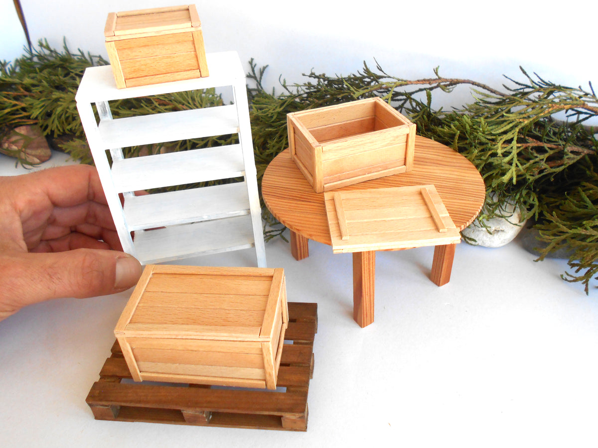 A miniature dollhouse furniture set featuring a light wood crate on a wooden pallet, a round table with a matching crate and tray, and a white shelving unit with a crate on top, shown with a hand holding the shelf for scale.