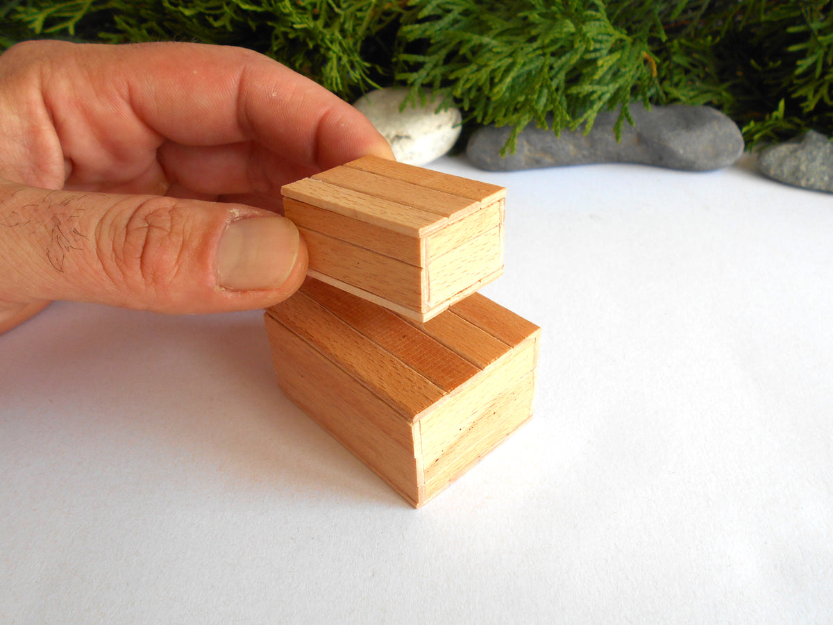 A hand holding a small wooden block above a larger matching wooden block on a white surface, with greenery and stones in the blurred background.