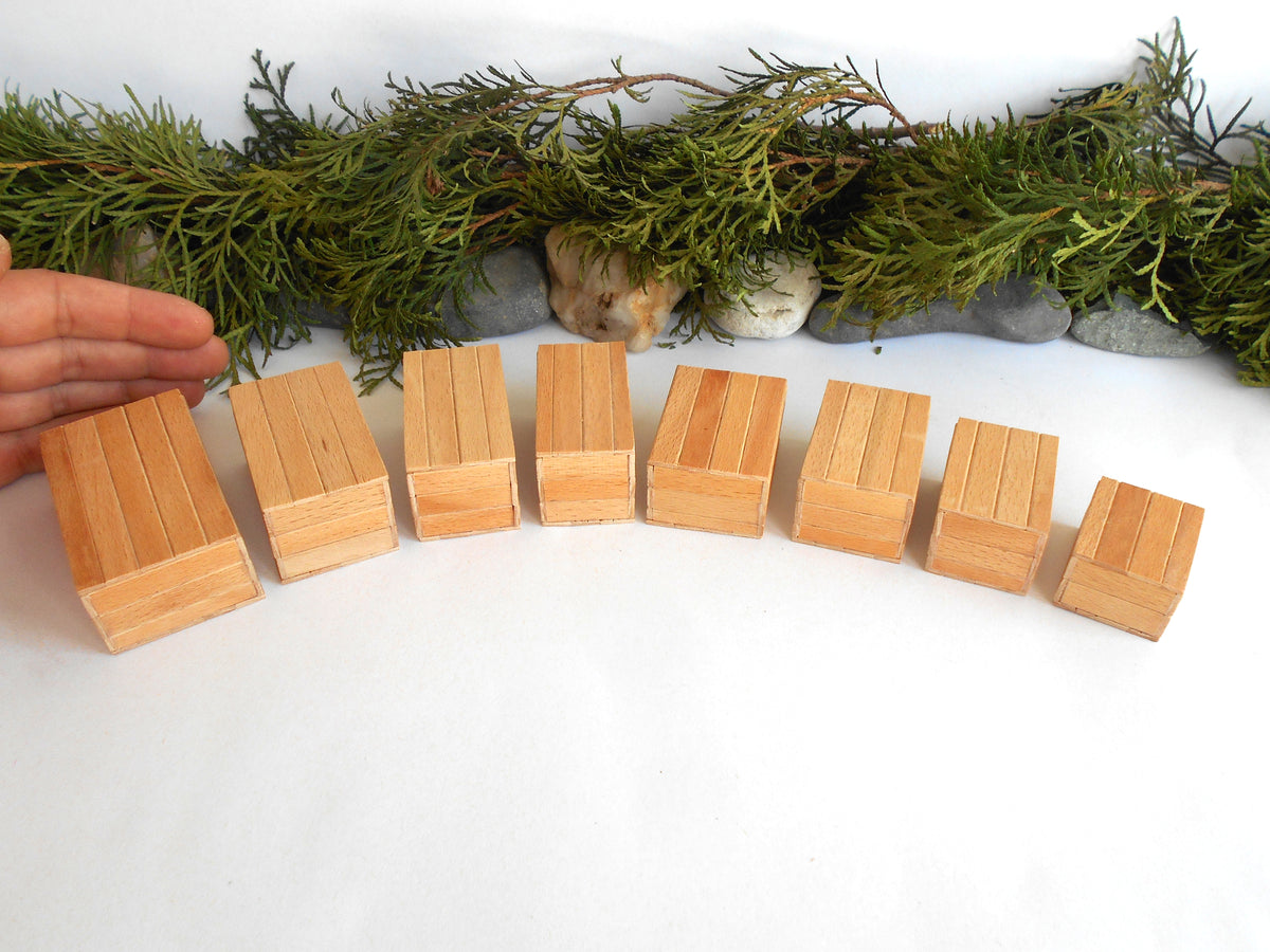A set of small natural wood boxes arranged in a curved row on a white surface, with a hand entering from the left and evergreen branches and stones behind them.
