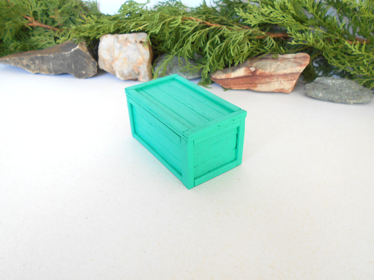 A miniature bright green wooden crate or storage box with a lid, photographed on a white surface with decorative greenery and stones in the background.