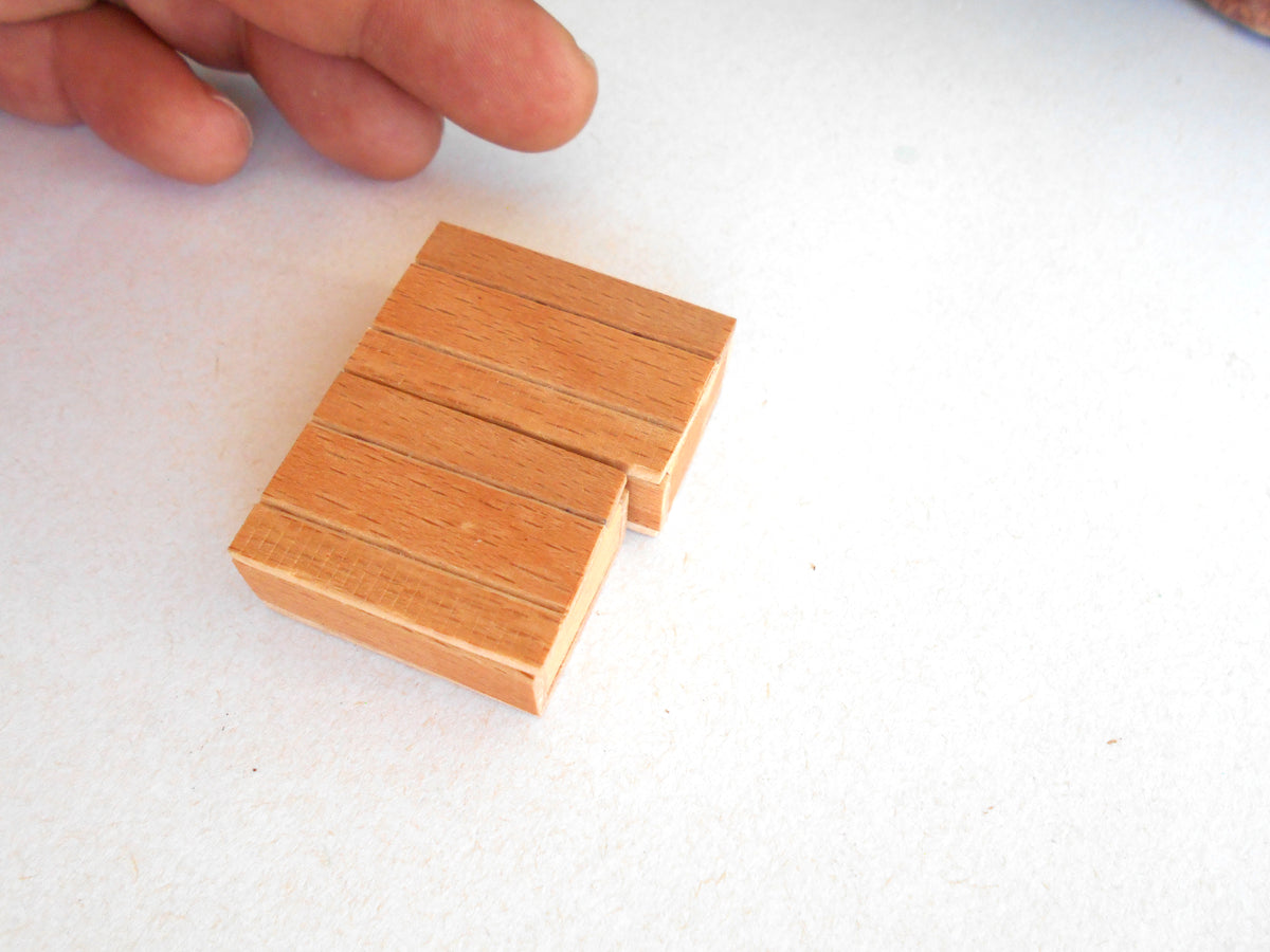 A small wooden miniature box  with a light natural wood finish, shown on a white surface with a person's fingers pointing at it from the top left.