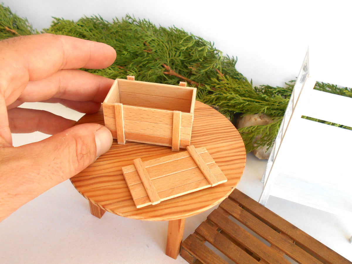 A hand holds a miniature wooden crate model next to a small round wooden table, with miniature greenery and a white slatted chair in the background.