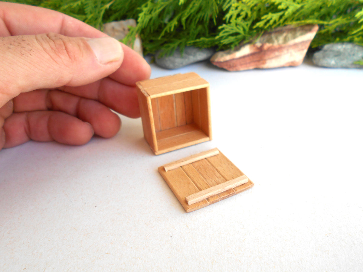 A miniature wooden crate or box with its lid placed beside it, held by a person's hand on a white surface with decorative greenery in the background.