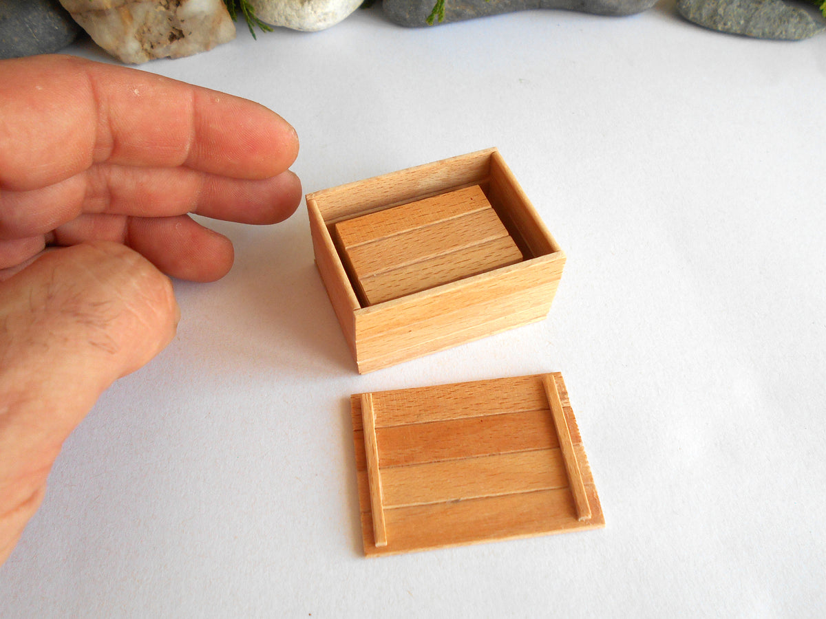 A miniature light wood box with its lid placed beside it on a white surface, with a person's hand visible for scale.